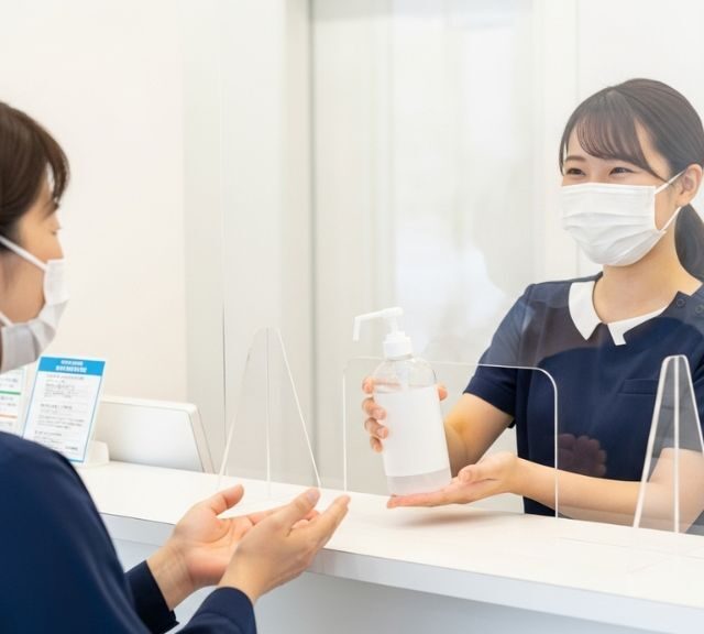 消毒液を感染防止アクリルボードごしに患者に渡すクリニックの受付スタッフ